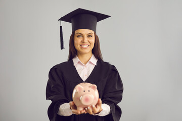 Student saving money to pay for postgraduate college or university education abroad. Happy woman in academic gown and mortarboard holding piggy bank and looking at camera standing on gray background