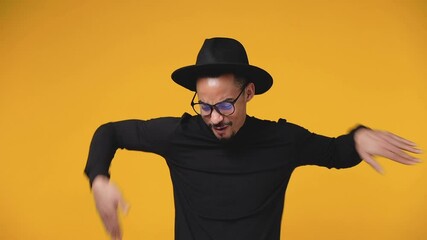 Wall Mural - Cheerful african american man 20s in black clothes hat eyeglasses isolated on yellow background studio. People lifestyle concept. Dancing clenching fists waving rising hands pointing fingers sing song