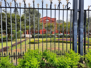 Canvas Print - Red building and the garden behind the gray gate with beautiful golden ornaments