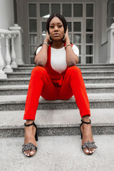 Wall Mural - Stylish young black woman with sexy lips in fashionable elegant clothes in stylish sandals posing sitting on stone vintage stair in city on summer day. Attractive fashion model black girl in red suit.