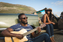 Diverse Couple Taking Roadside Break On Sunny Day Beside Convertible Car The Man Playing Guitar