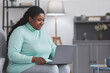 © Seventyfour - Portrait of curvy African American woman using laptop and smiling while enjoying work from home sitting on couch in minimal grey interior, copy space