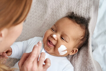 caucasian mother applying healthy skin care moisturizing cream on cute adorable funny african americ