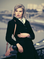 Wall Mural - Fashion blond woman in black coat with beige leather handbag leaning on railing