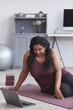 Vertical portrait of curvy African American woman working out at home while sitting on yoga mat and watching online training video