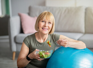 Wall Mural - Exercise and healthy diet concept. Senior woman with fitball eating fresh vegetable salad at home