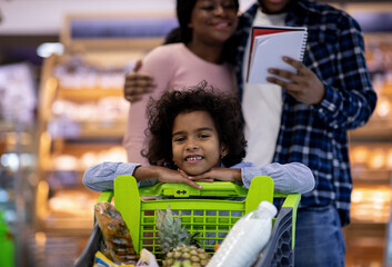 Adorable black girl with full shopping cart and her parents checking grocery list at modern supermarket