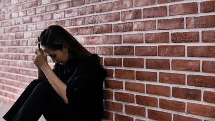 Wall Mural - Christian Asian woman is sitting against a wall, feeling sad, holding a cross and praying to God.