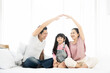 © Monster Ztudio - Happy asian family sitting on bed in bedroom together and making the home sign. Mom and dad making roof figure with hands arms over heads