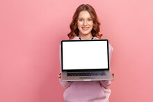Portrait Of Happy Smiling Curly Haired Teenage Girl In Hoodie Showing Empty Laptop Screen On Camera, Place For Advertisement. Indoor Studio Shot Isolated On Pink Background