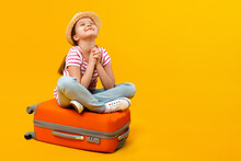 The Child Sits On A Suitcase And, Closing His Eyes, Dreams Of Travel, Adventure, Vacation. Little Girl On A Yellow Background.
