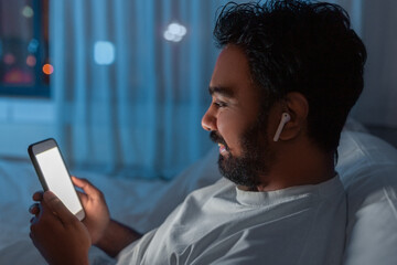 Poster - technology, internet, communication and people concept - happy smiling young indian man with smartphone and earphones listening to music in bed at night