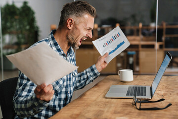 Wall Mural - Furious grey man screaming while working with laptop and papers