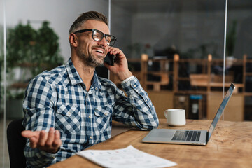 Wall Mural - Smiling grey man talking on mobile phone while working with laptop
