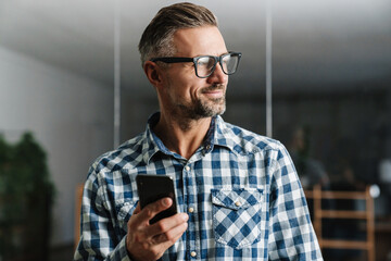 Wall Mural - Happy white-haired man in eyeglasses smiling while using mobile phone