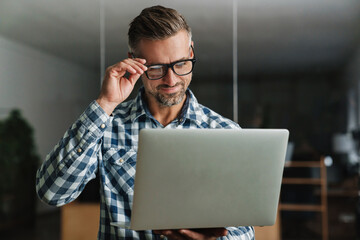 Wall Mural - Smiling handsome man working with laptop while standing in office