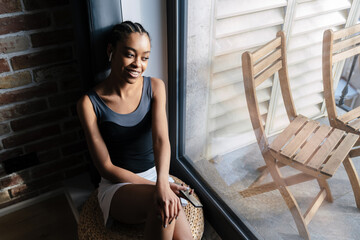 Wall Mural - Happy african american girl in earphones using cellphone while sitting at window