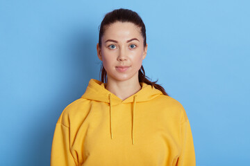young serious young woman has serious expression, wears casual yellow hoodie, poses against blue bac