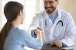 © fizkes - Smiling young handsome male doctor in eyeglasses and uniform shaking hands with female client, celebrating signing medical insurance contract or welcoming patient at checkup meeting in clinic office.
