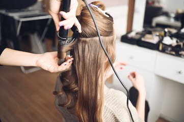 Wall Mural - Hair stylist prepares beautiful young woman for event, makes curls hairstyle with a curling iron for client in beauty salon. Long beautiful light brown natural hair.