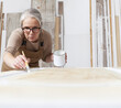 © amedeoemaja - wood crafts, woman artisan carpenter painting with brush and paint jar white the door in workshop, wearing overall and eyeglasses, interior designer, restoration, diy and handmade works concept