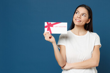 Wall Mural - Young dreamful pensive wistful latin woman in basic white t-shirt hold gift voucher flyer mock up do winner gesture gesture clench fist celebrating isolated on dark blue background studio portrait