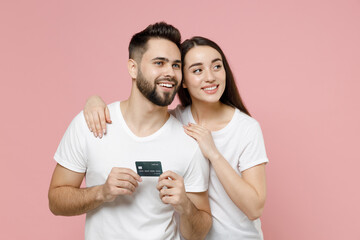 Canvas Print - Young lovely couple two friends bearded man brunette woman in white basic blank print design t-shirts hold in hands mockup of credit bank card isolated on pastel pink color background studio portrait.