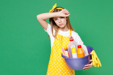 Exhausted tired little kid girl housewife 12-13 in yellow apron put hand on head hold basin detergent bottles washing cleansers doing housework isolated on green wall background. Housekeeping concept.