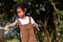 Little African American Curly Hair Girl In Casual Clothing Holding Bubble Wand Blowing Bubbles Playing Alone At Outdoor.