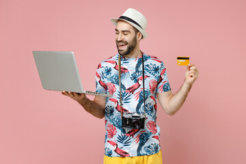 Funny traveler tourist man in summer clothes hat using laptop pc computer booking hotel hold credit bank card isolated on pink background. Passenger traveling on weekends. Air flight journey concept.