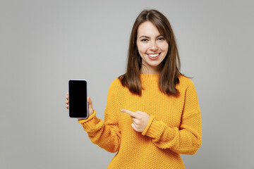 Young smiling attractive beautiful caucasian woman 20s in casual knitted yellow sweater point index finger mobile phone with blank screen workspace area isolated on grey background studio portrait