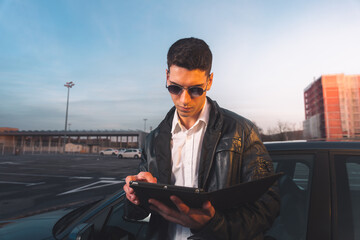 Young caucasian using a digital tablet with a sports car behind in a parking lot.