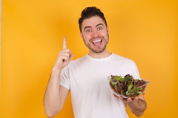 Poster - young handsome Caucasian man holding a salad bowl against yellow wall says: wow how exciting it is, has amazed expression, indicates something. One hand on her chest and pointing with other hand.