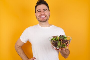 Wall Mural - Studio shot of cheerful young handsome Caucasian man holding a salad bowl against yellow wall keeps hand on hip, smiles broadly.