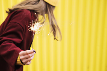 Unknown woman in burgundy coat and holding a burning sparkler in her hands against the background of bright yellow wall.