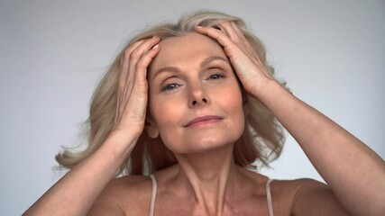 happy 50s middle aged woman with flying blond hair touching facial skin looking at camera pampering 