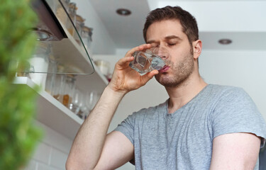 Unshaven man drinks water from a glass in the kitchen.