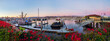 A panorama of a calm harbor of ships under a pink sunset with red and pink bougainvillea plants in the foreground.  There are multiple ships in the harbor, with white docks visible. 