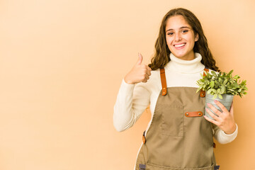 Wall Mural - Young caucasian gardener woman holding a plant isolated smiling and raising thumb up