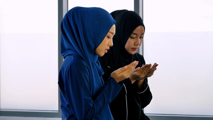 Two Asian Muslim young women in traditional hijab are praying glorify Allah and practicing the Islamic faith in mosque.