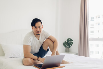 Asian man is sitting on the bed and working on his laptop.