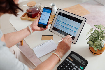 Hands of senior woman checking utility bill on screen tablet computer and paying with banking application on smartphone