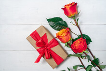 Three Red And Yellow Roses And A Gift Box On A White Wooden Background