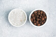 Coarse salt and black peppercorns in white ceramic bowls on gray concrete background, copy space top view.