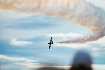 Aerial demonstration of a military airplane making a turn in the sky as it leaves a smoke trail, blurred heads of people watching