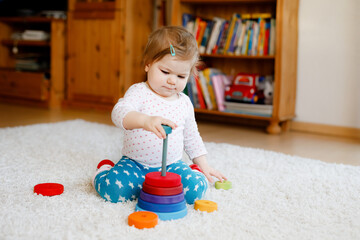 Adorable cute beautiful little baby girl playing with educational wooden toys at home or nursery. Toddler with colorful stack pyramid and music toy. Happy healthy child having fun with different toys