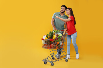 Wall Mural - Young couple with shopping cart full of groceries on yellow background