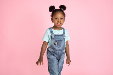 Wall Mural - Cute little African preschool girl, wearing casual jeans overalls and t-shirt, posing to camera on isolated pink studio background. People, kids, studio portraits