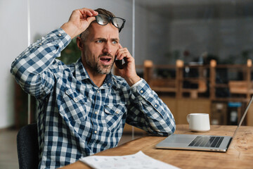 Wall Mural - Shocked grey man talking on mobile phone while working with laptop