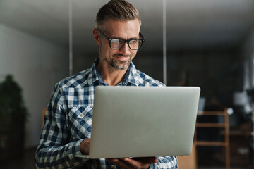 Wall Mural - Smiling handsome man working with laptop while standing in office